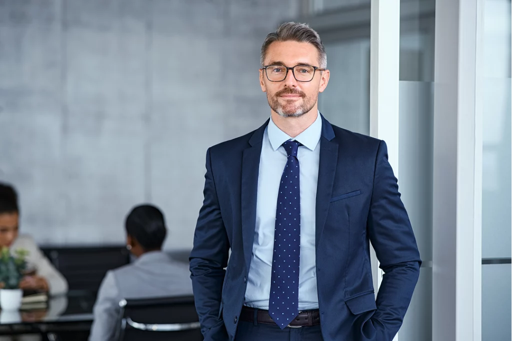 Geschäftsmann im Anzug steht selbstbewusst im Büro, im Hintergrund arbeiten Kollegen an einem Tisch.