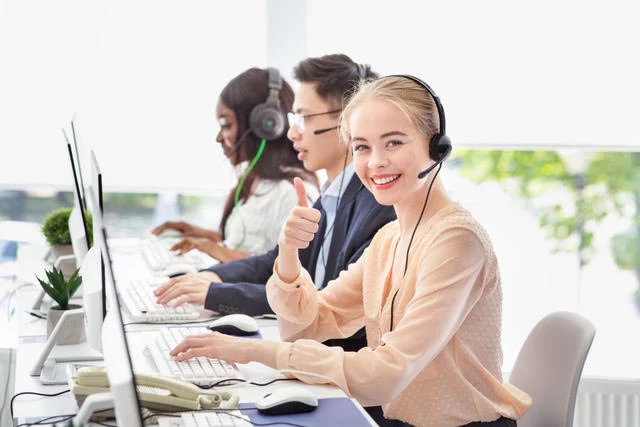 Freundliches Service-Team mit Headsets im Büro, im Vordergrund eine lächelnde Mitarbeiterin mit Daumen hoch.