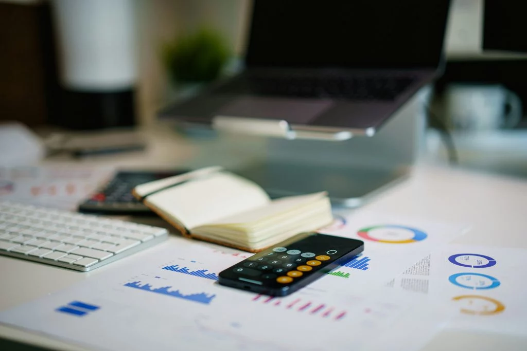 Schreibtisch mit Laptop, Tastatur, Notizbuch, Taschenrechner und Diagrammen für Analyse, Planung und Finanzübersicht.