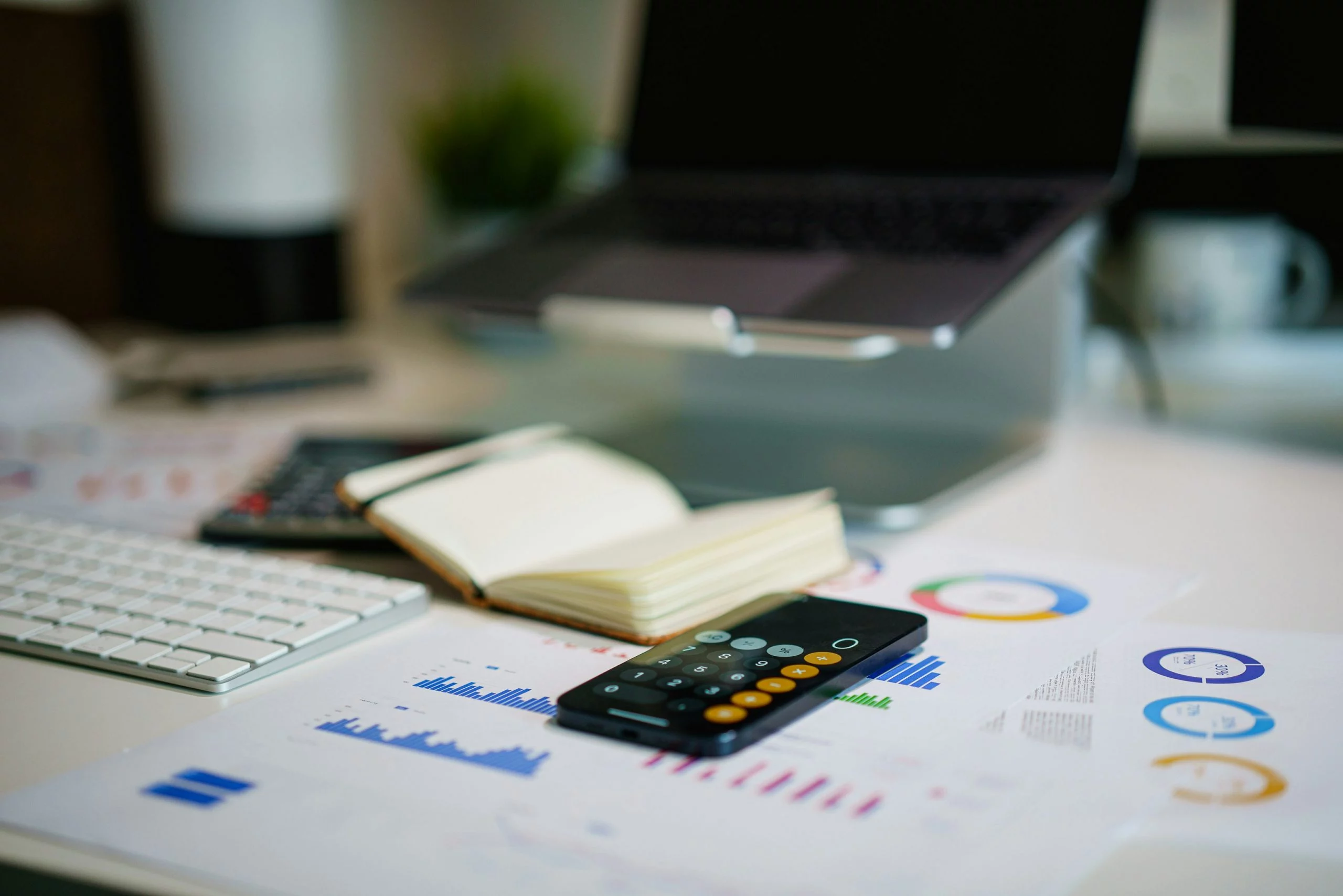 Schreibtisch mit Laptop, Tastatur, Notizbuch, Taschenrechner und Diagrammen für Analyse, Planung und Finanzübersicht.