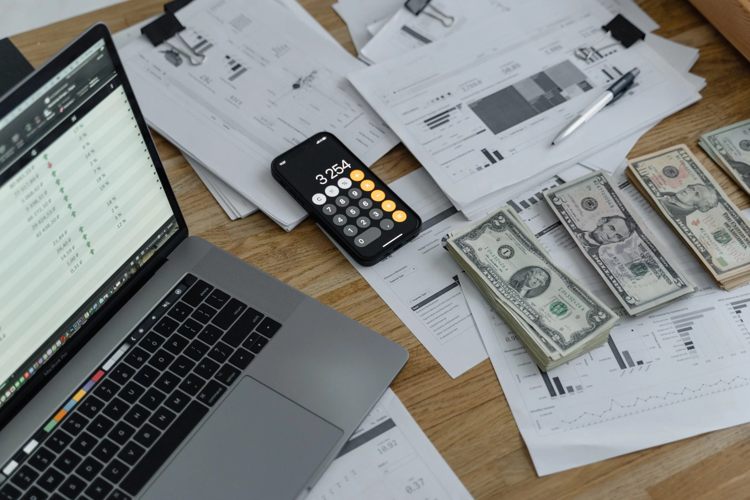Laptop, Smartphone mit Rechner-App, Unterlagen und US-Dollar-Scheine auf einem Schreibtisch.